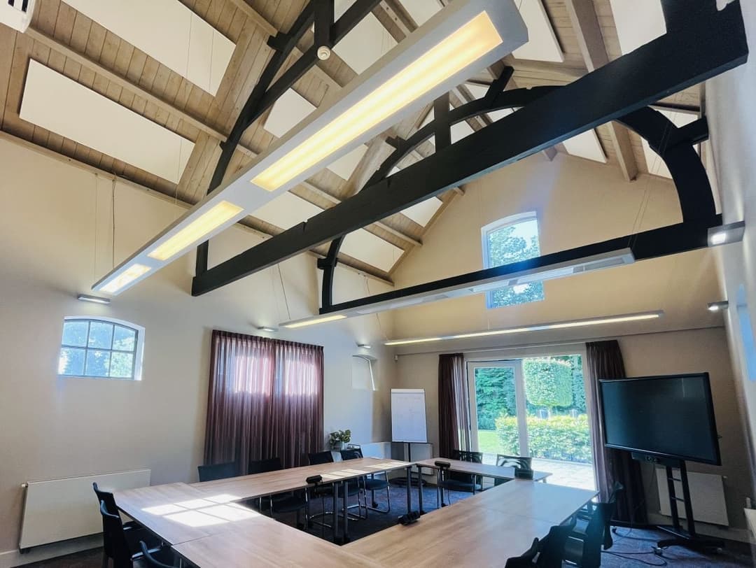 Achttien EASYceiling Plus akoestische panelen in de kleur White Frost zijn gemonteerd tegen een houten plafond in een vergaderzaal. De witte panelen zorgen voor geluidsabsorptie en verbeteren de akoestiek van de ruimte.